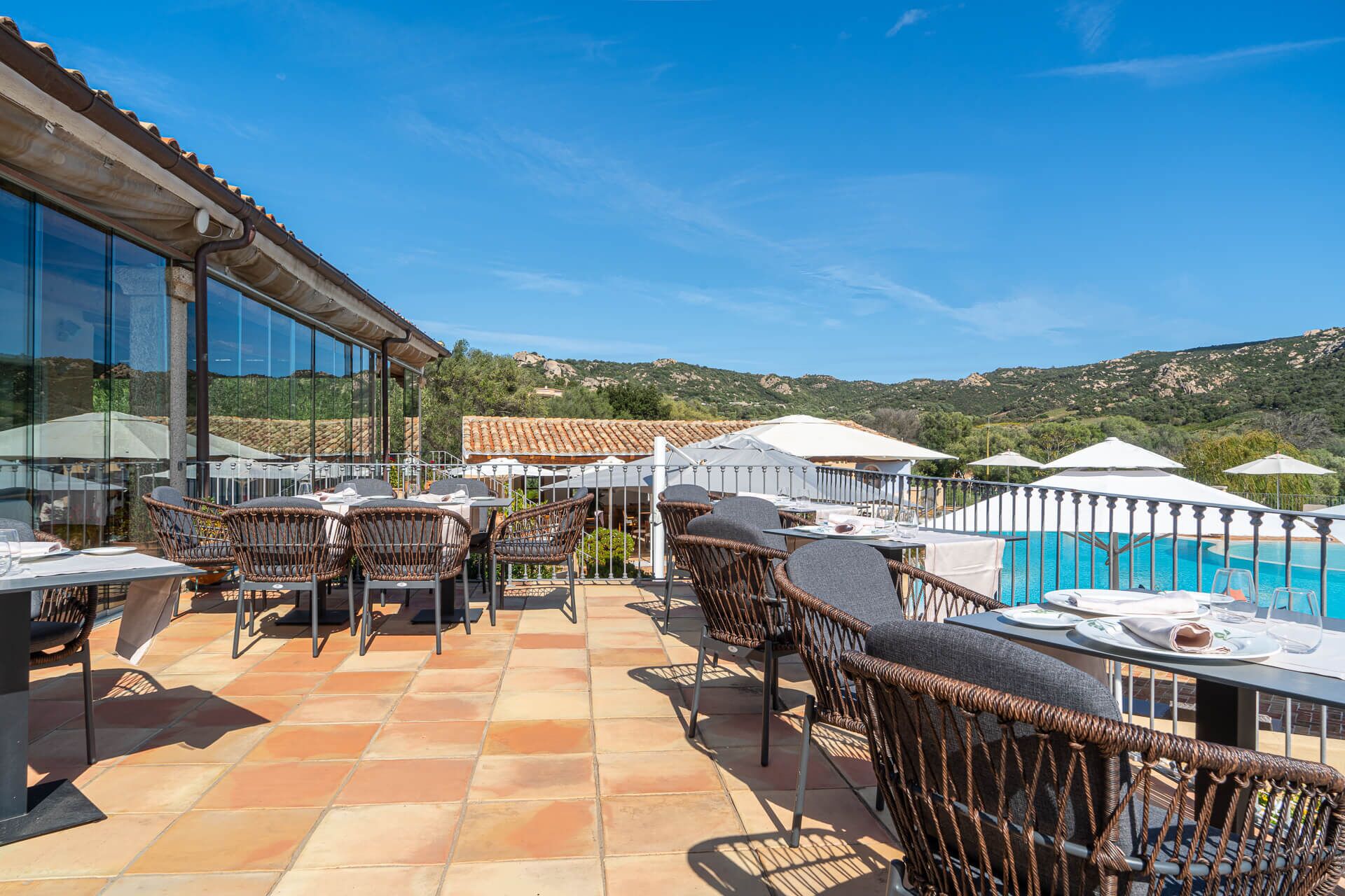 An outdoor dining area with wicker chairs and tables, overlooking a swimming pool and a mountainous landscape.