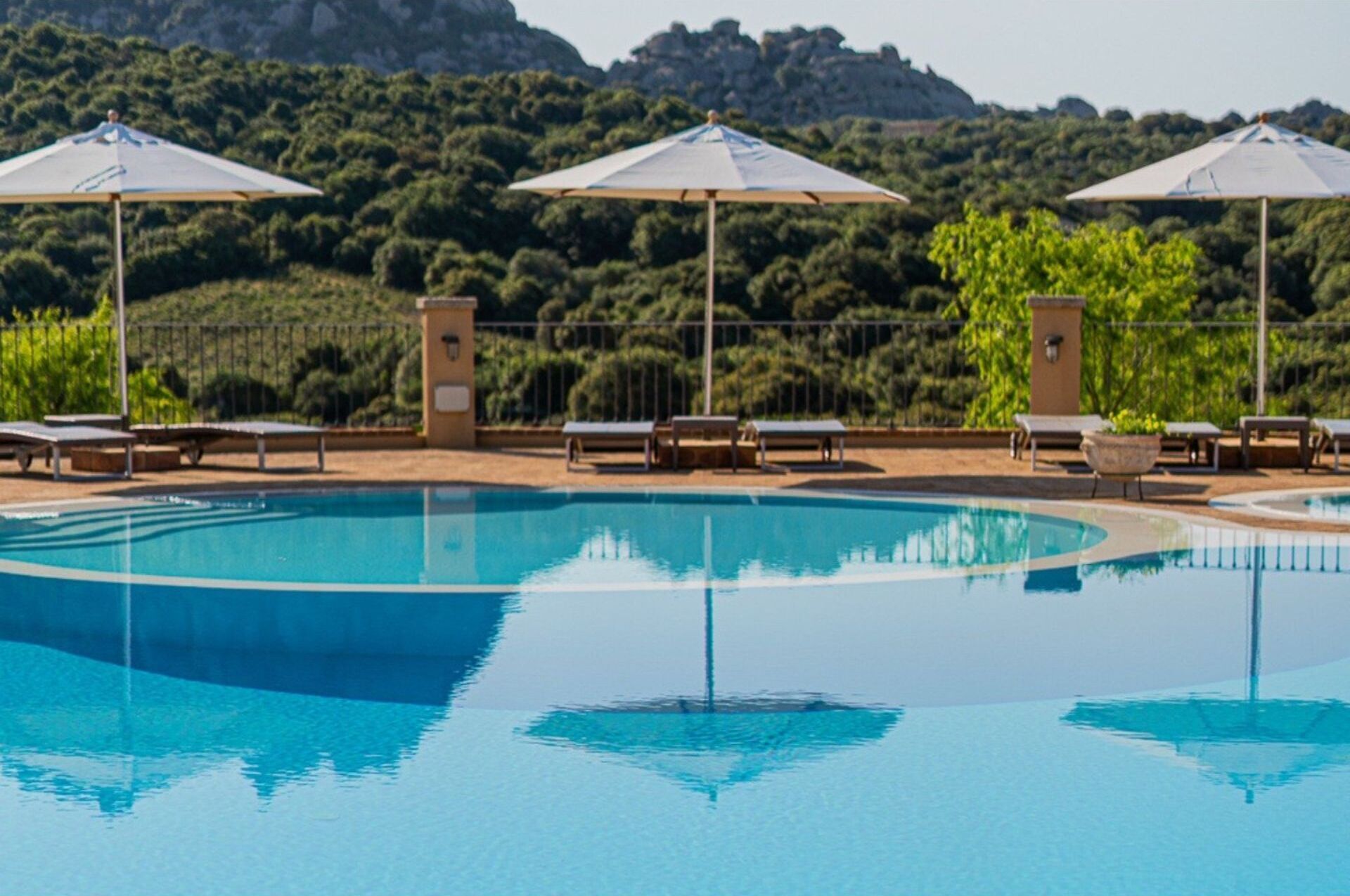 A pool area with lounge chairs and umbrellas, surrounded by greenery and hills.