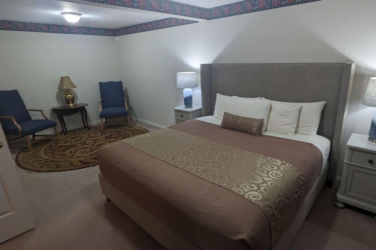 Ein Hotelzimmer mit einem großen Bett, zwei Stühlen, einem Nachttisch mit Lampe und Teppichboden.