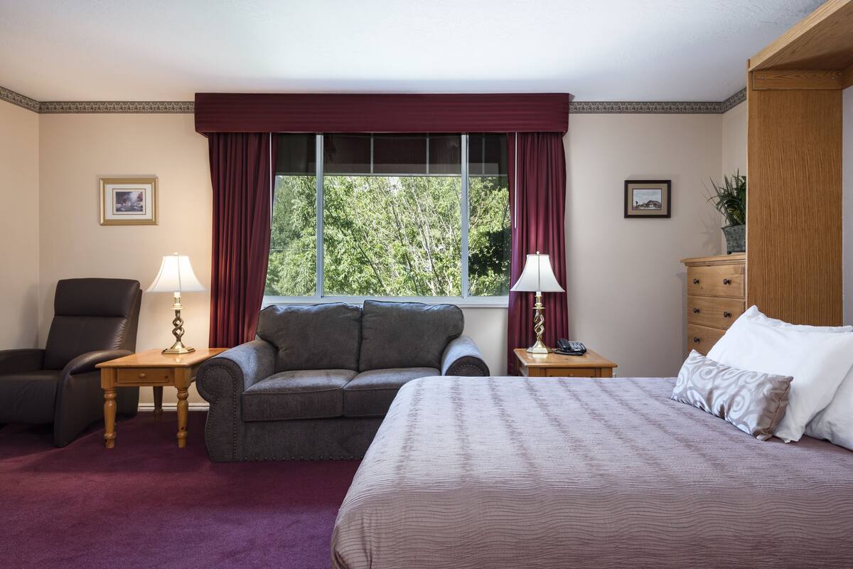 Ein Hotelzimmer mit einem Bett, einem Sofa, einem Sessel, zwei Nachttischen mit Lampen, einer Holzkommode und einem Fenster mit Vorhängen.