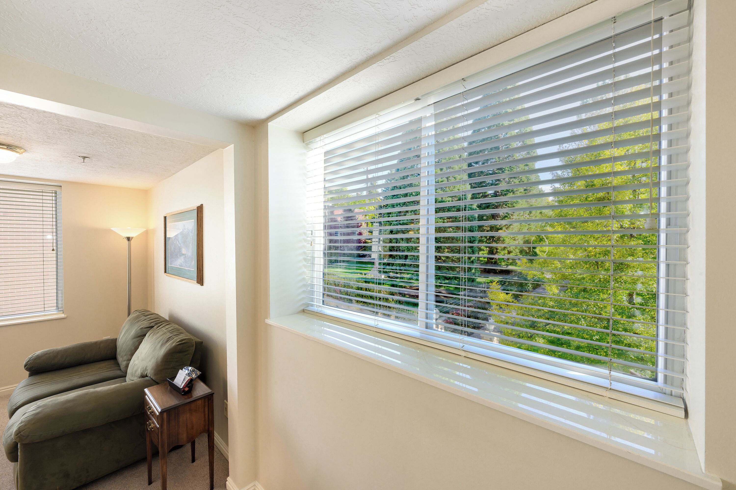 A room with a large window with blinds, a green sofa, a small table, and a lamp.