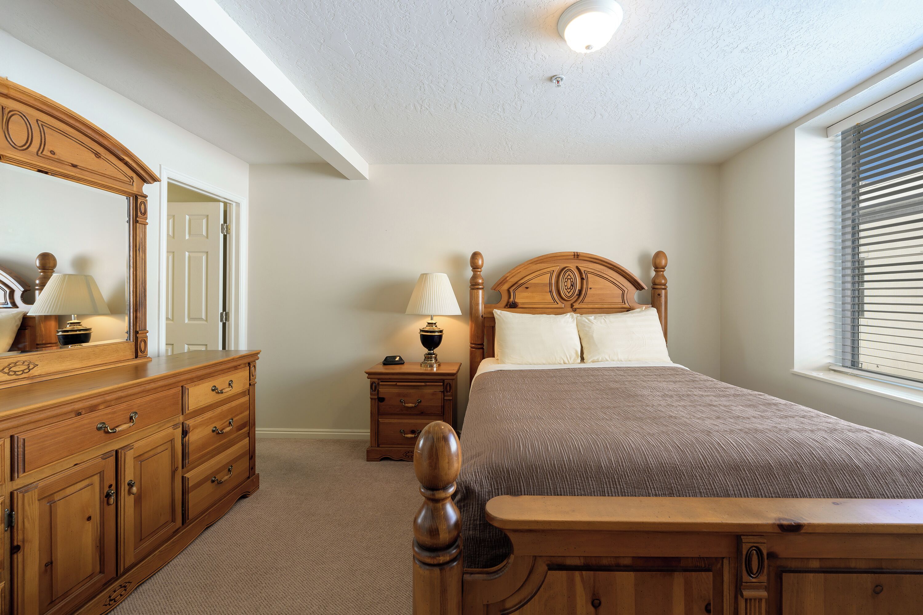 Quarto com cama de madeira, mesa de cabeceira com abajur, cômoda com gavetas e um espelho grande.