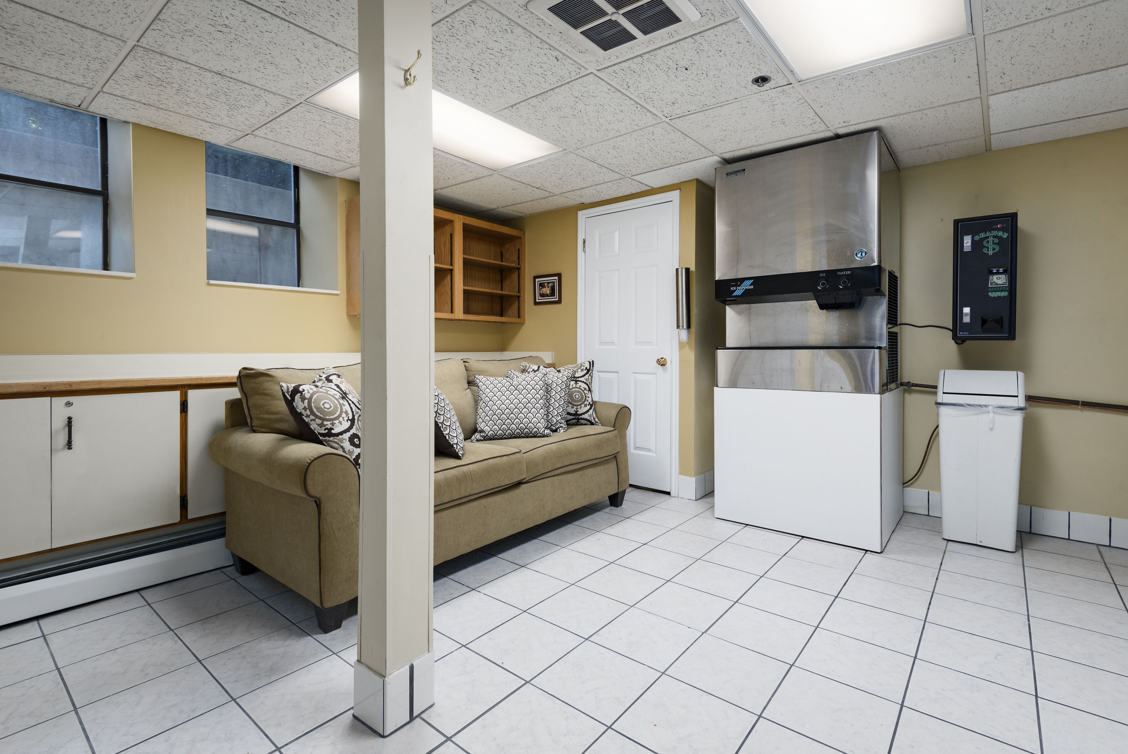 A kitchenette with a refrigerator, stove, and microwave. There is a beige sofa with patterned cushions. The room has a tiled floor and a white door.