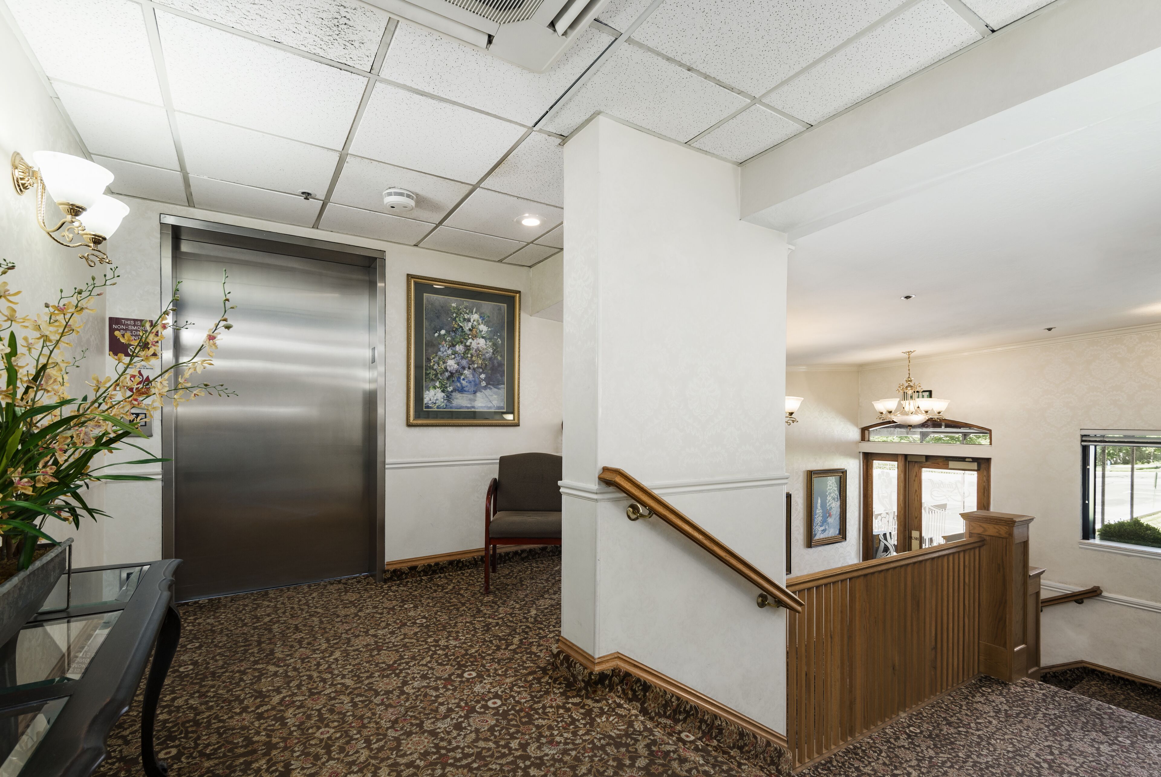 A hotel lobby with a carpeted floor, a reception desk, a chair, a staircase, and a framed picture on the wall.