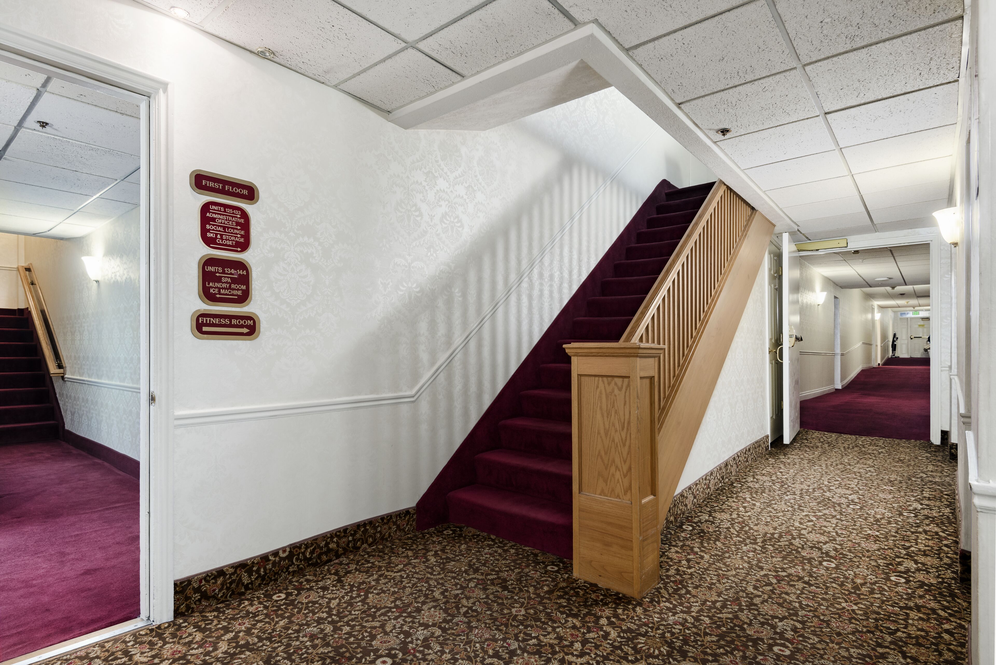 Pasillo de hotel con piso alfombrado, una escalera de madera y señales indicativas para el primer piso, incluyendo un gimnasio.