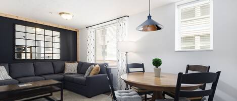 A modern living room with a dark sofa, a wooden coffee table, a dining table with chairs, and a pendant light.