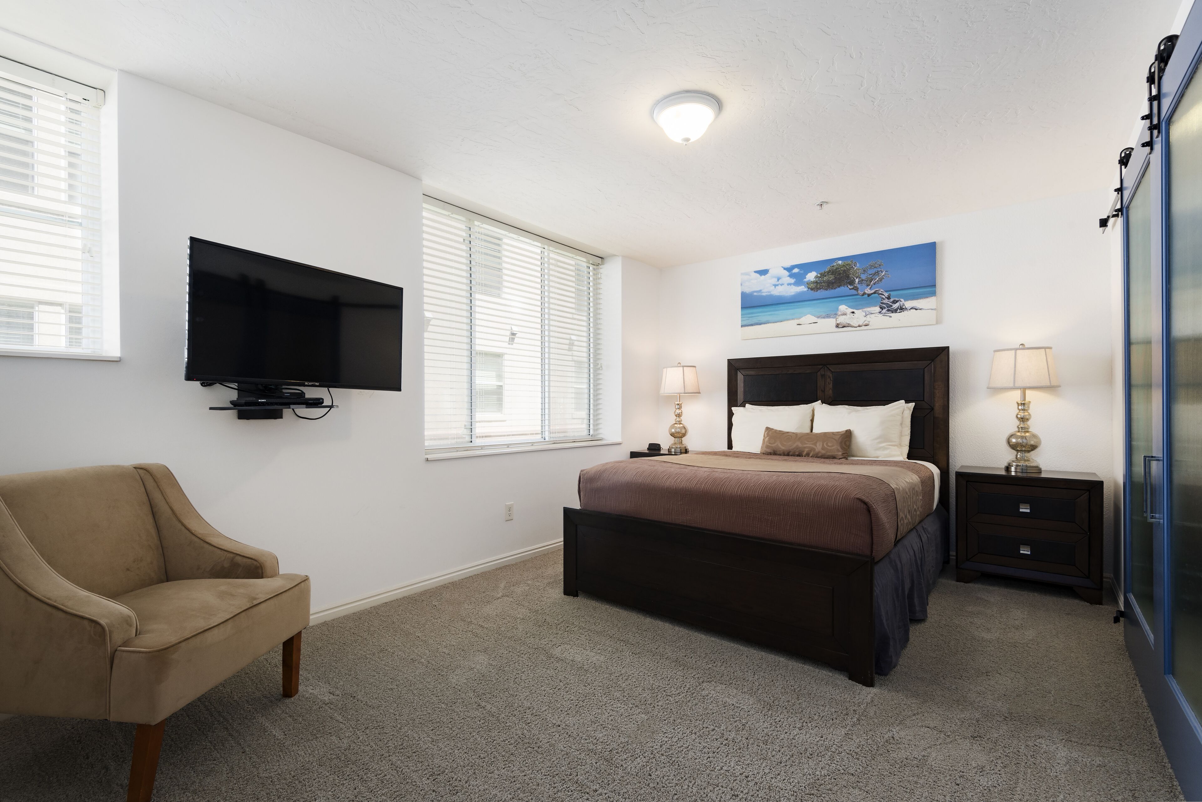 Un dormitorio con una cama, un televisor montado en la pared, una silla y un cuadro de una playa.