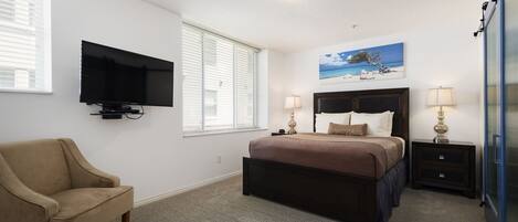 Un dormitorio con una cama, un televisor montado en la pared, una silla y un cuadro de una playa.