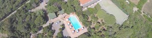 An aerial view of a property with a rectangular swimming pool surrounded by lounge chairs and umbrellas, a tennis court, and dense greenery. - Hotel Club Mezzaluna (Carloforte)