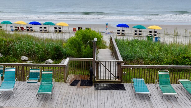 A wooden deck with green lounge chairs and a view of the beach with colorful umbrellas. - Ocean Forest Colony (Myrtle Beach)