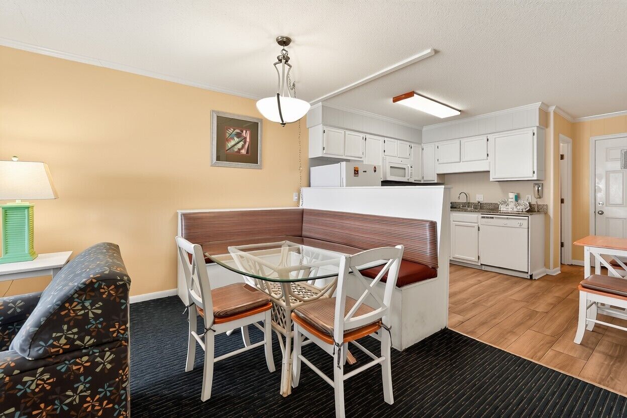 A compact kitchen with white cabinets and a microwave, a dining area with a glass table and chairs, and a sofa with a patterned cushion.