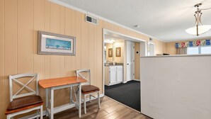 A room with a wooden dining table, two chairs, a framed picture on the wall, and a kitchen area in the background. - Ocean Forest Colony (Myrtle Beach)