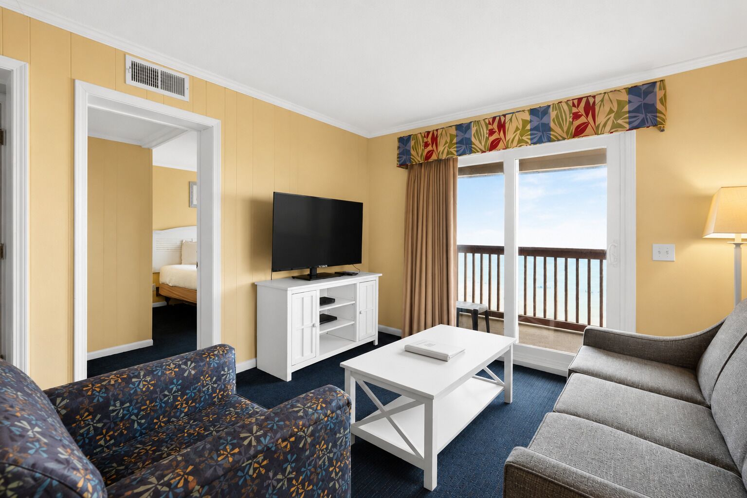 A hotel room with a bed, a TV on a white stand, a coffee table, a sofa, and a balcony with a view.
