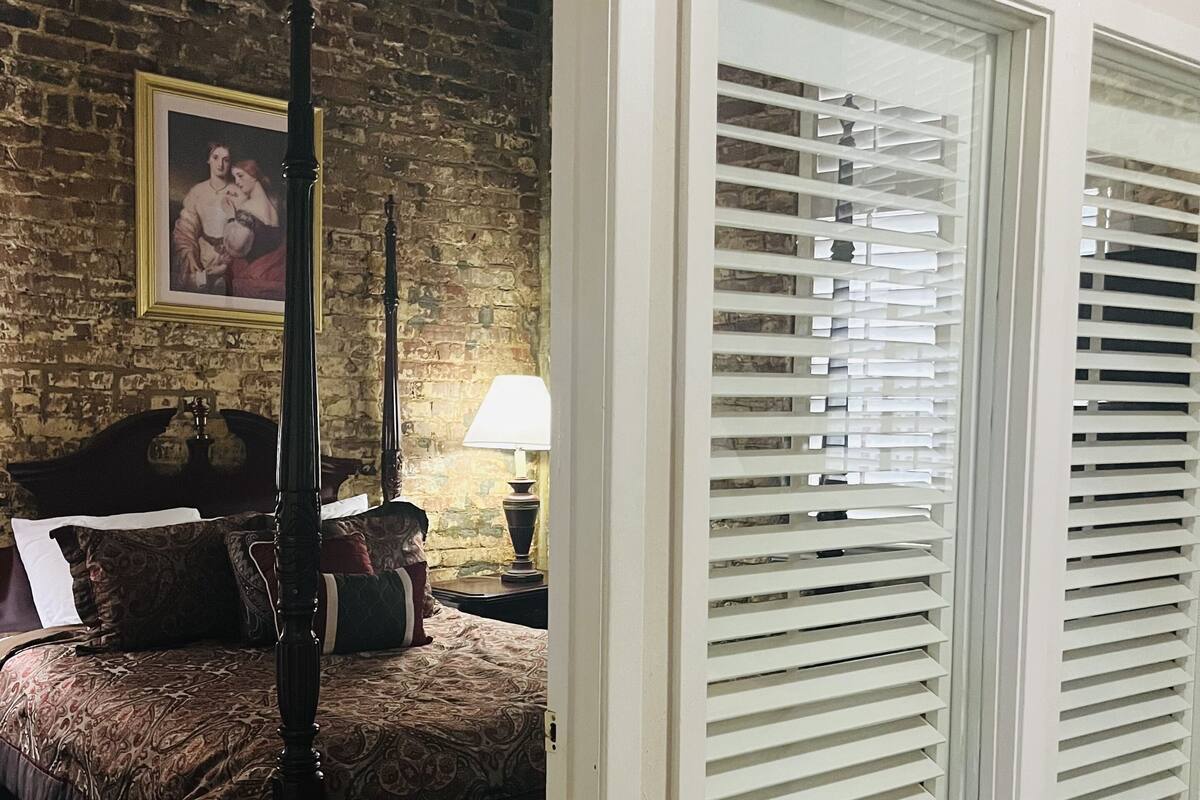 Un dormitorio con cama con dosel, pared de ladrillo, un cuadro enmarcado, mesita de noche con lámpara y contraventanas blancas.