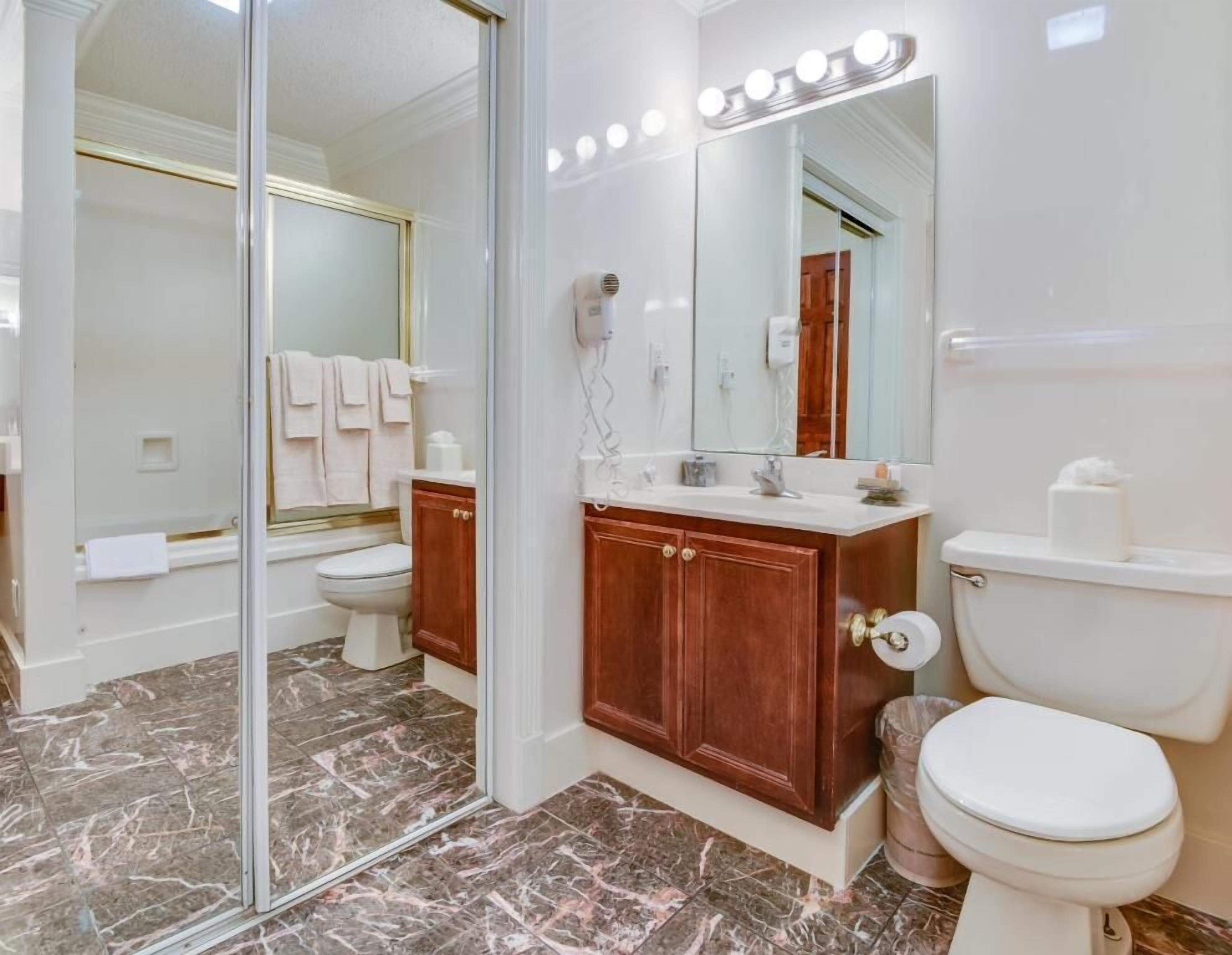 Une salle de bain dotée d’une douche à parois de verre, d’un meuble-lavabo avec un miroir et des luminaires, de toilettes et d’un revêtement de sol en marbre.