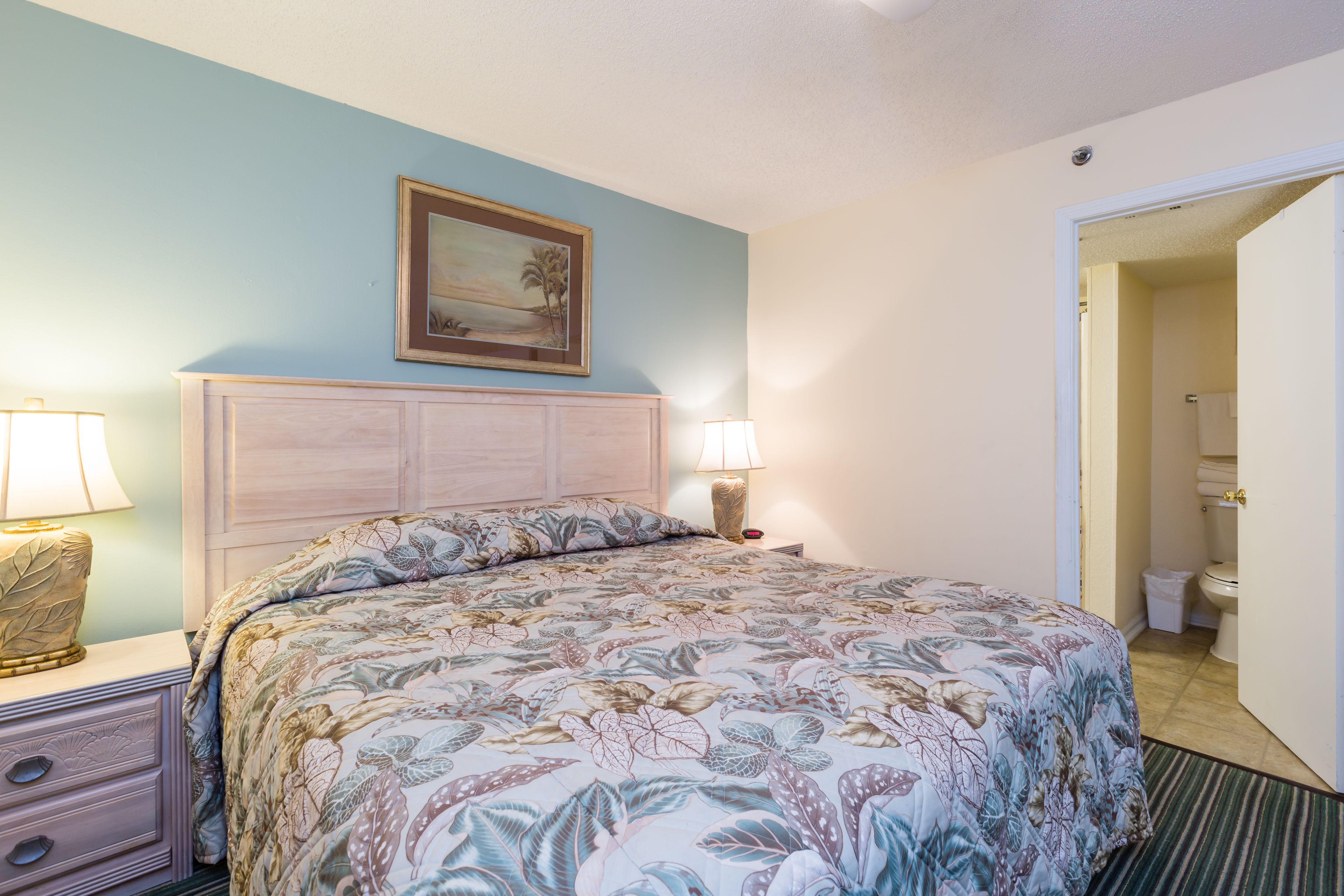 Une chambre à coucher avec une couvre-lit à motifs floraux, une table de chevet avec une lampe, une commode et un tableau accroché au mur.