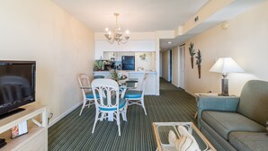 A compact living space with a TV, dining table, chairs, a sofa, a lamp, and a kitchen area in the background. - Windy Shores II (North Myrtle Beach)