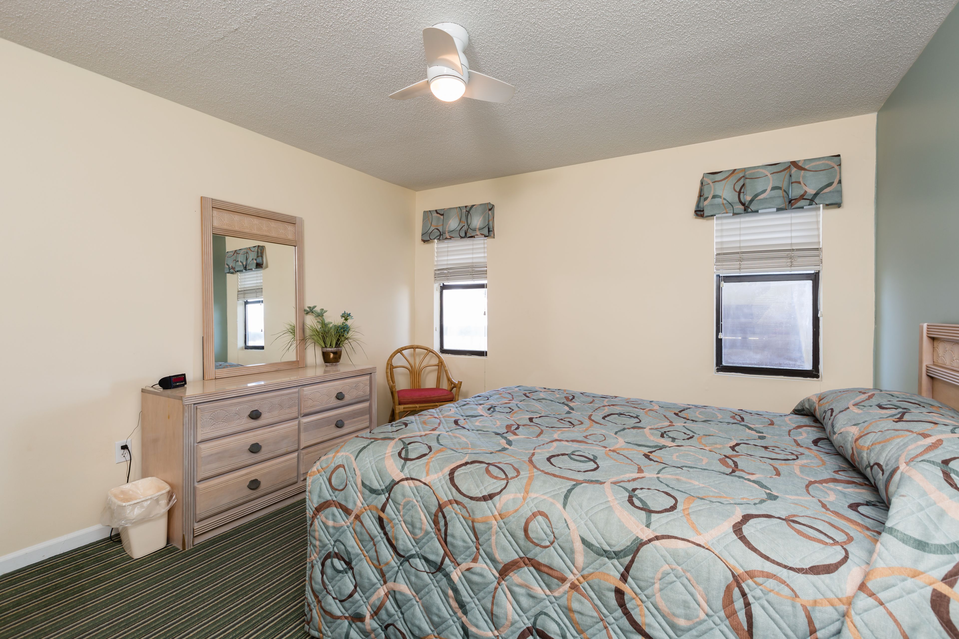 Un dormitorio con cama, cómoda, espejo, ventanas y ventilador de techo.