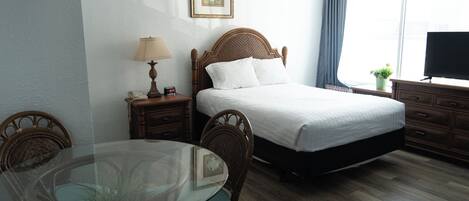 Quarto de hotel com cama, mesa de cabeceira com luminária, televisão, uma pequena planta e mesa de jantar com cadeiras.