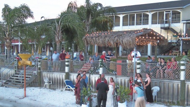 Uma cerimônia de casamento na praia, com um gazebo de palha e convidados sentados em cadeiras.