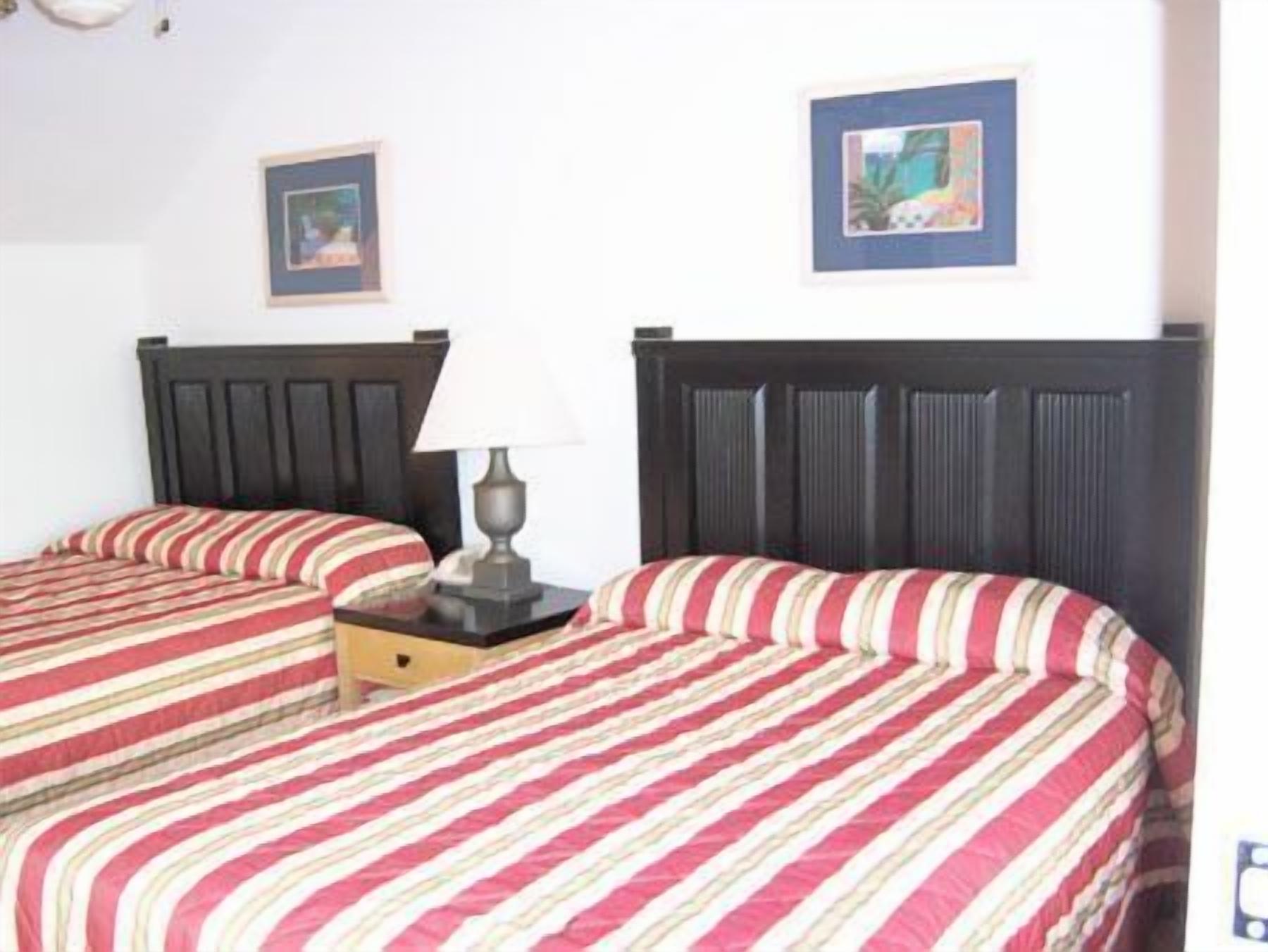 A hotel room with two beds, each with a dark headboard and striped bedding. There is a nightstand with a lamp between the beds.