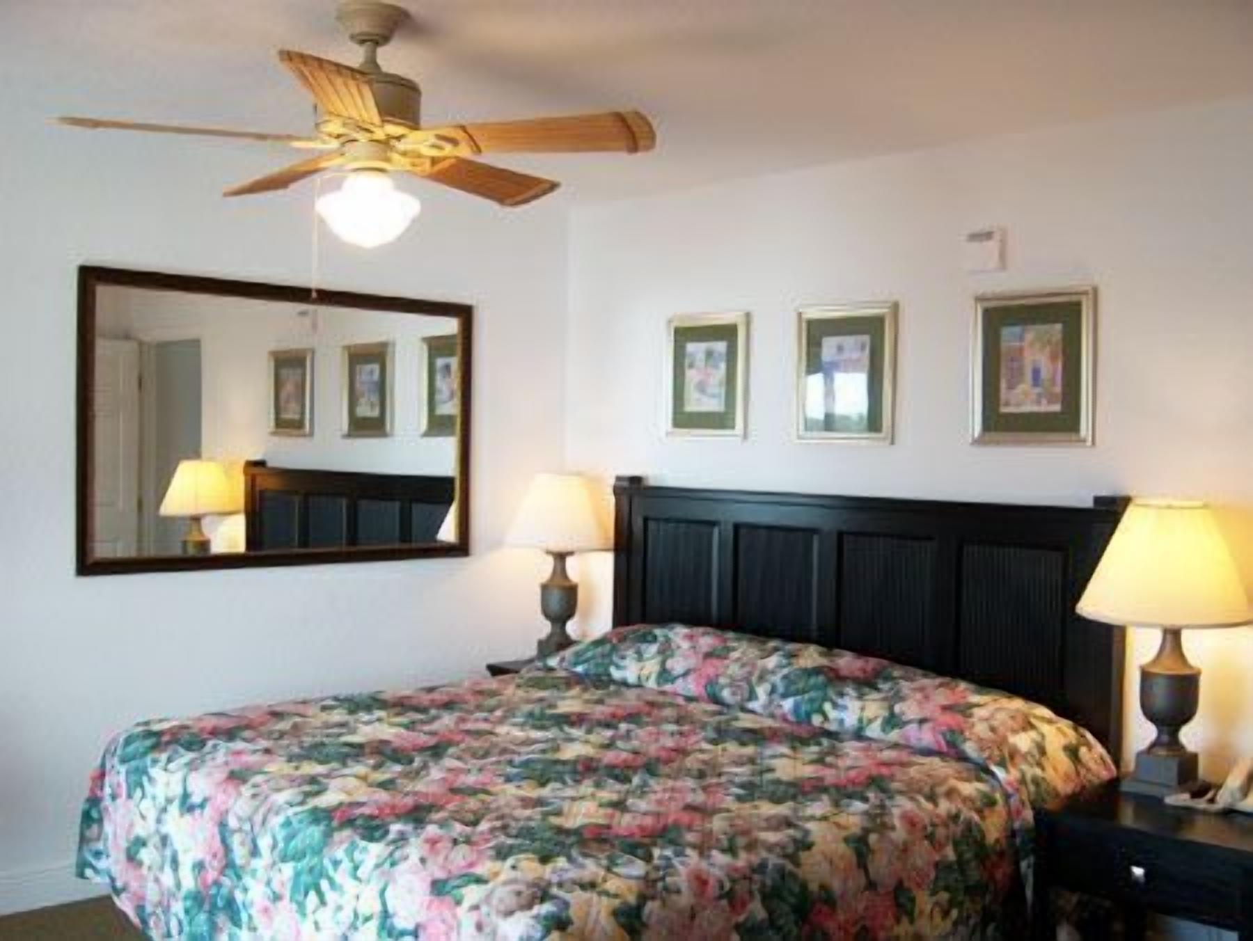 Une chambre à coucher comprenant un lit, un ventilateur de plafond, un grand miroir et deux lampes fixées au mur.