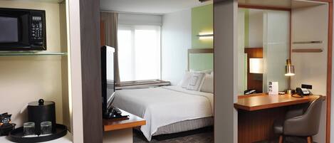 A hotel room with a bed, desk, chair, and a microwave on a shelf.