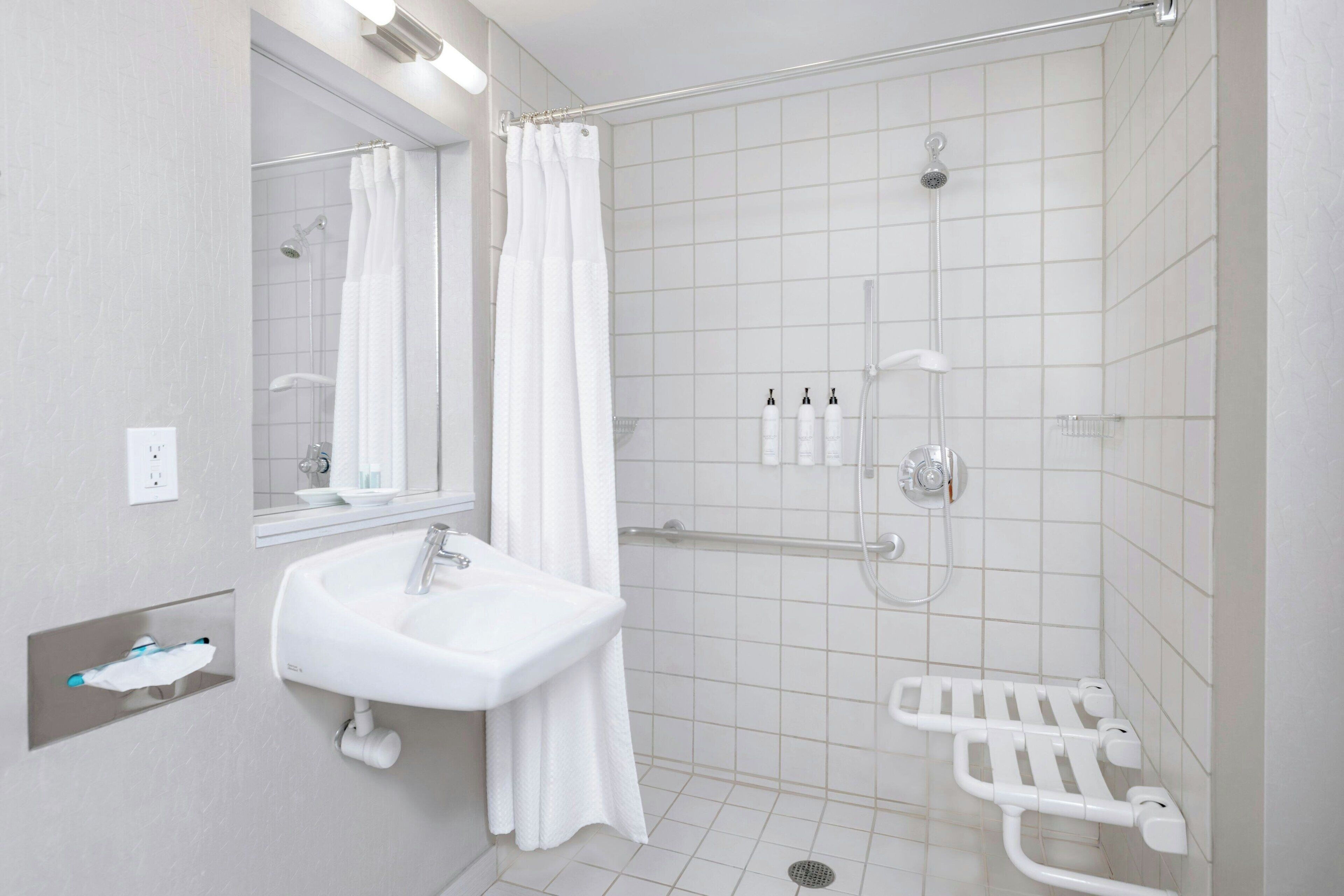 Un baño moderno con lavamanos blanco, espejo, ducha y barras de apoyo.