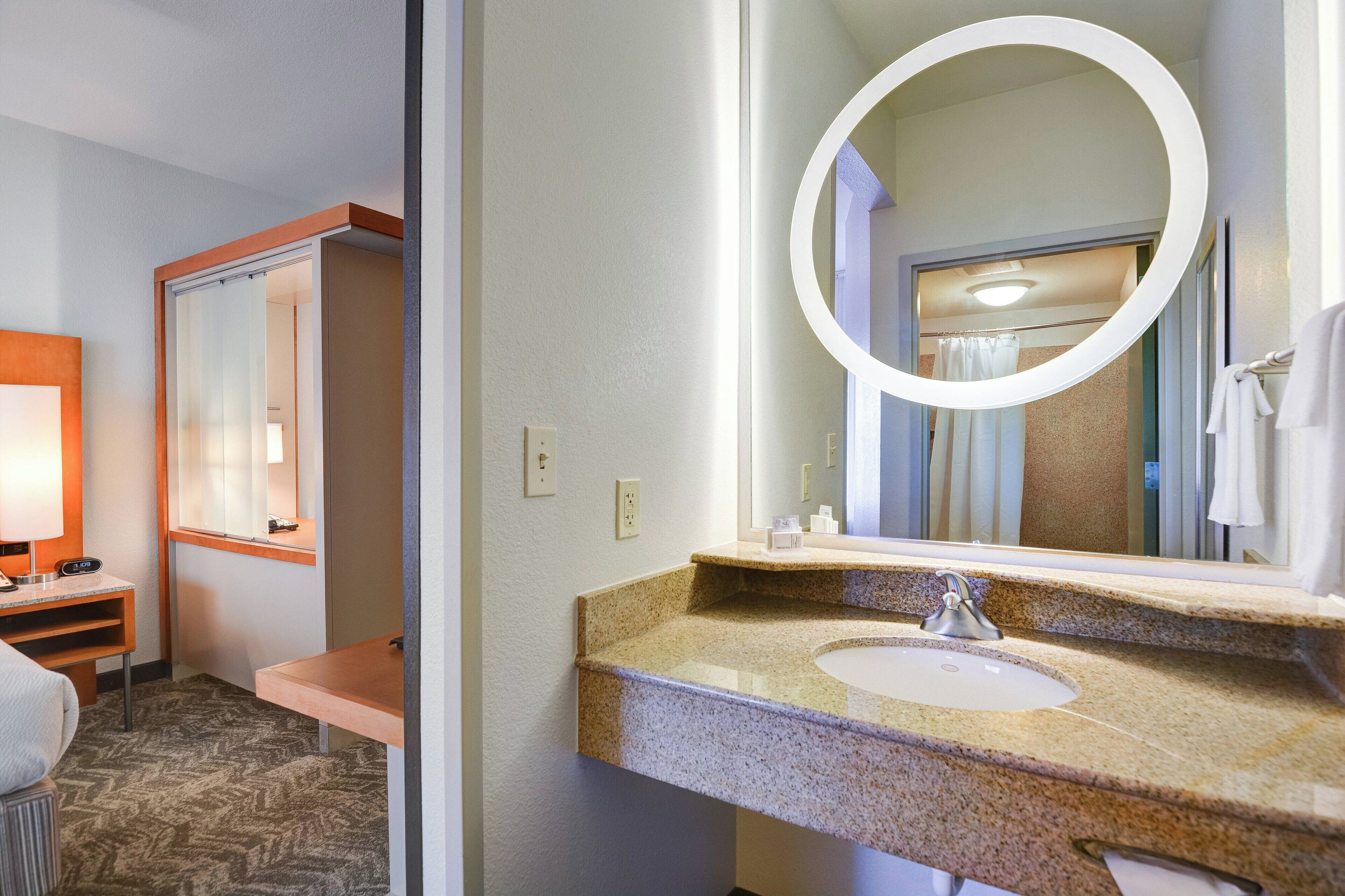 A hotel bathroom with a granite sink, a round mirror, a towel rack, and a shower visible through the glass door.