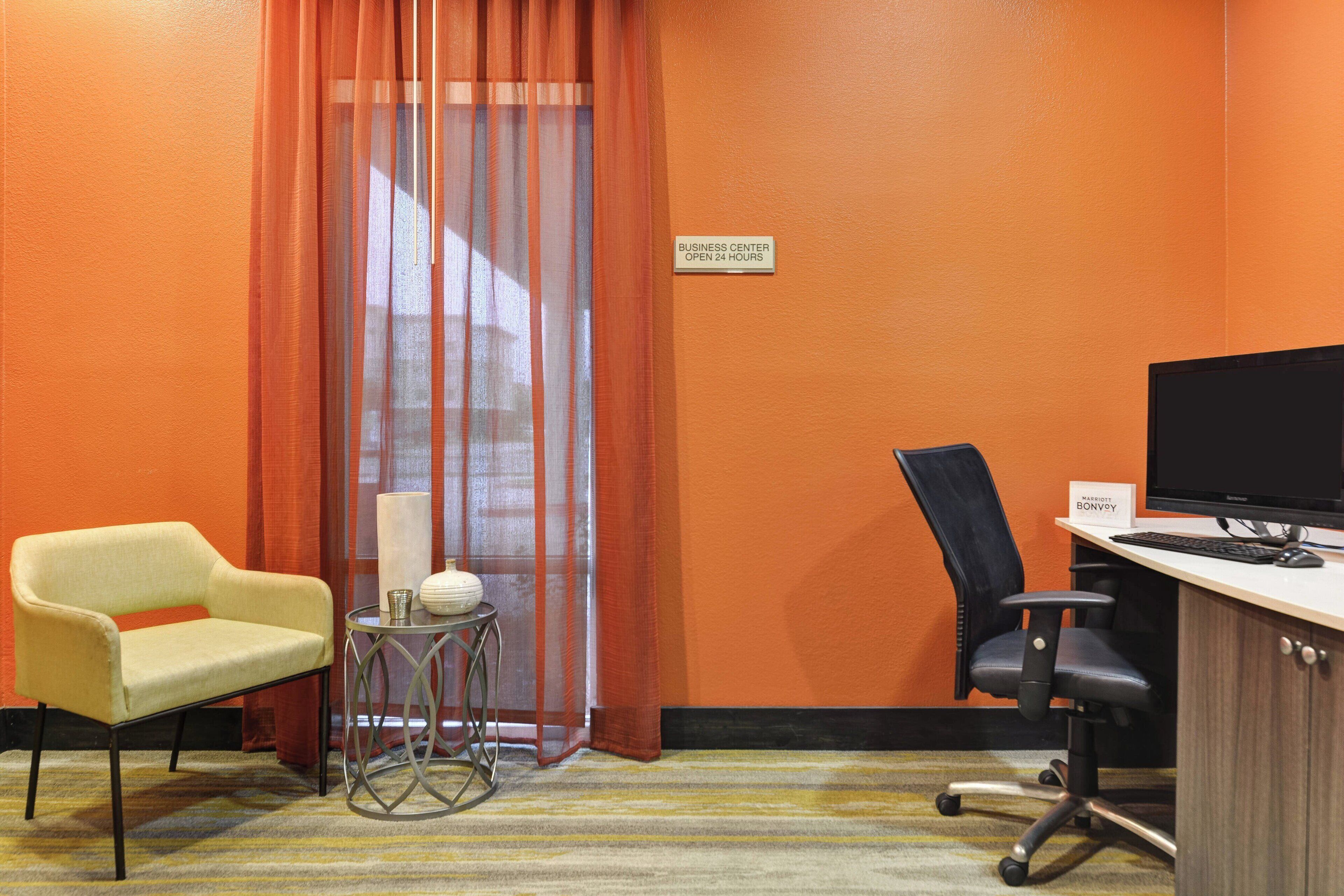 A hotel room with an orange wall, a desk with a computer, a black office chair, a yellow armchair, and a small table with a vase.