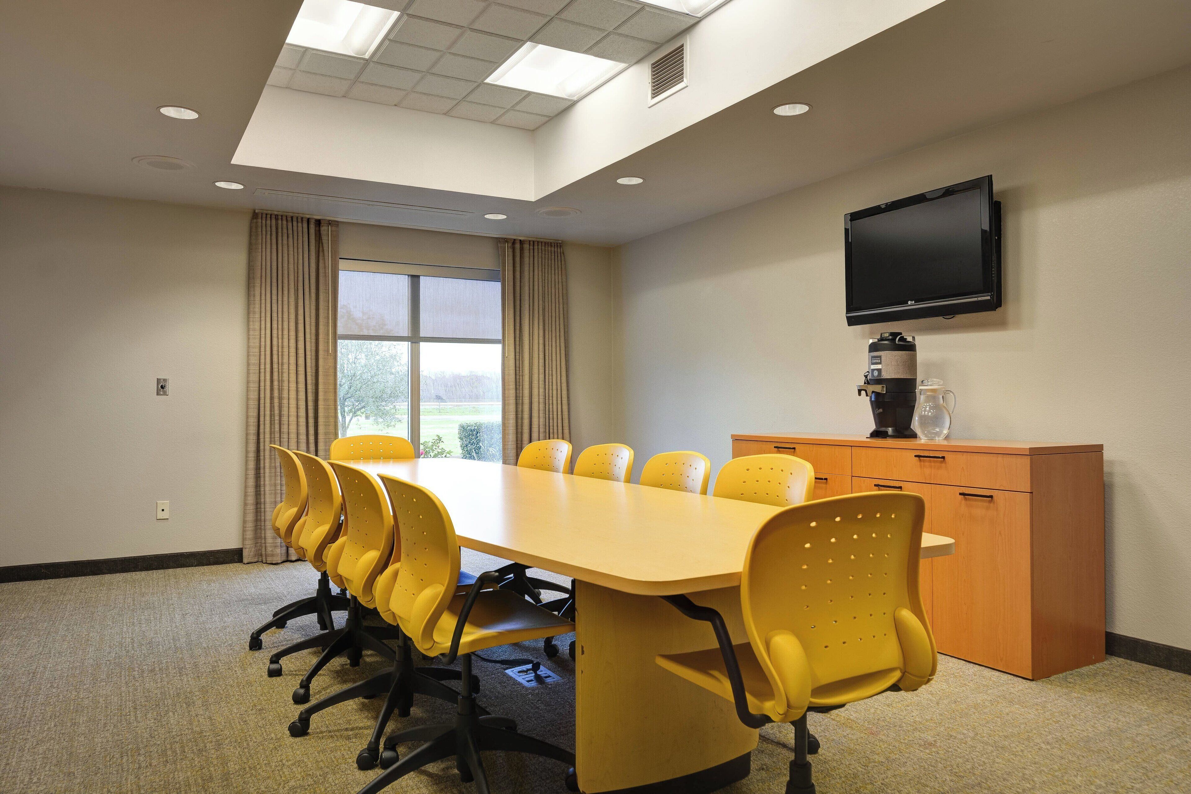 A conference room with a long table, yellow chairs, a television on the wall, and a coffee machine.