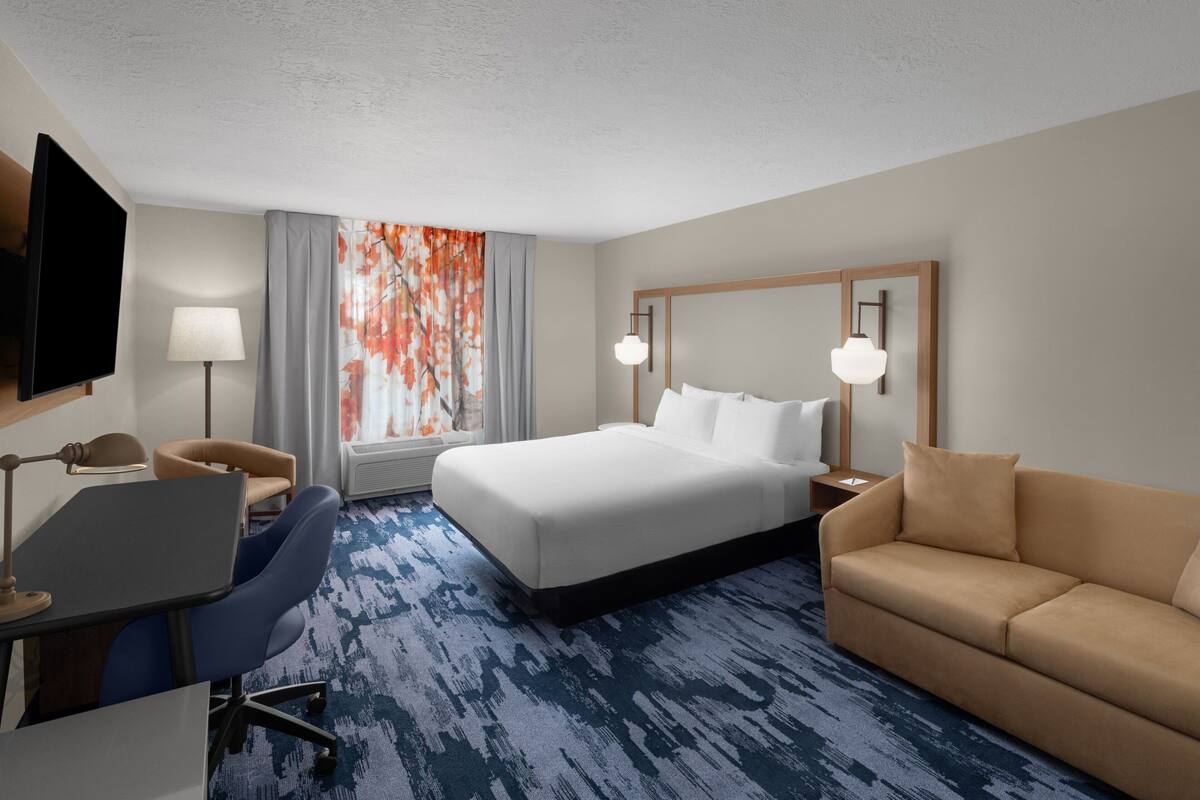 A hotel room with a large bed, a desk with a chair, a sofa, a TV, and a window with curtains.
