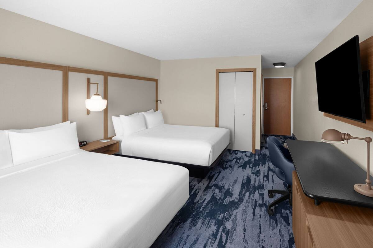 A hotel room with two beds, a desk, a chair, a TV, and a lamp.