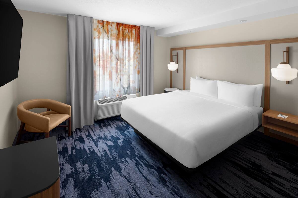 A hotel room with a large bed, a desk, a chair, a window with curtains, and a wall-mounted lamp.