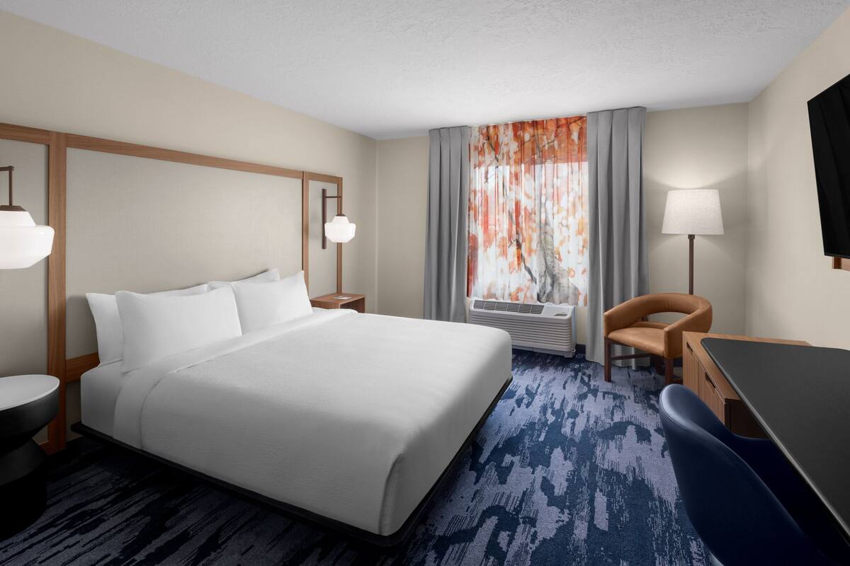 A hotel room with a large bed, a desk, a chair, a lamp, and a window with curtains.