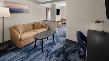A hotel room with a beige sofa, a small table, a desk with a lamp, a chair, a TV, and a painting on the wall.