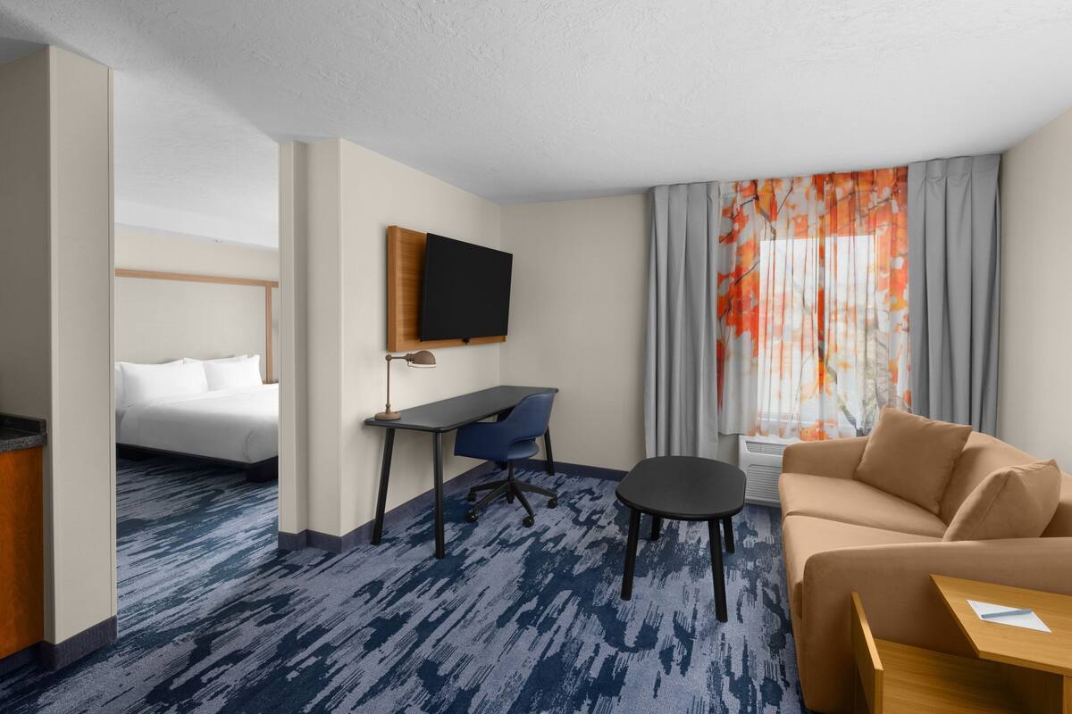 A hotel room with a bed, a desk with a chair, a sofa, a TV mounted on the wall, and a window with curtains.