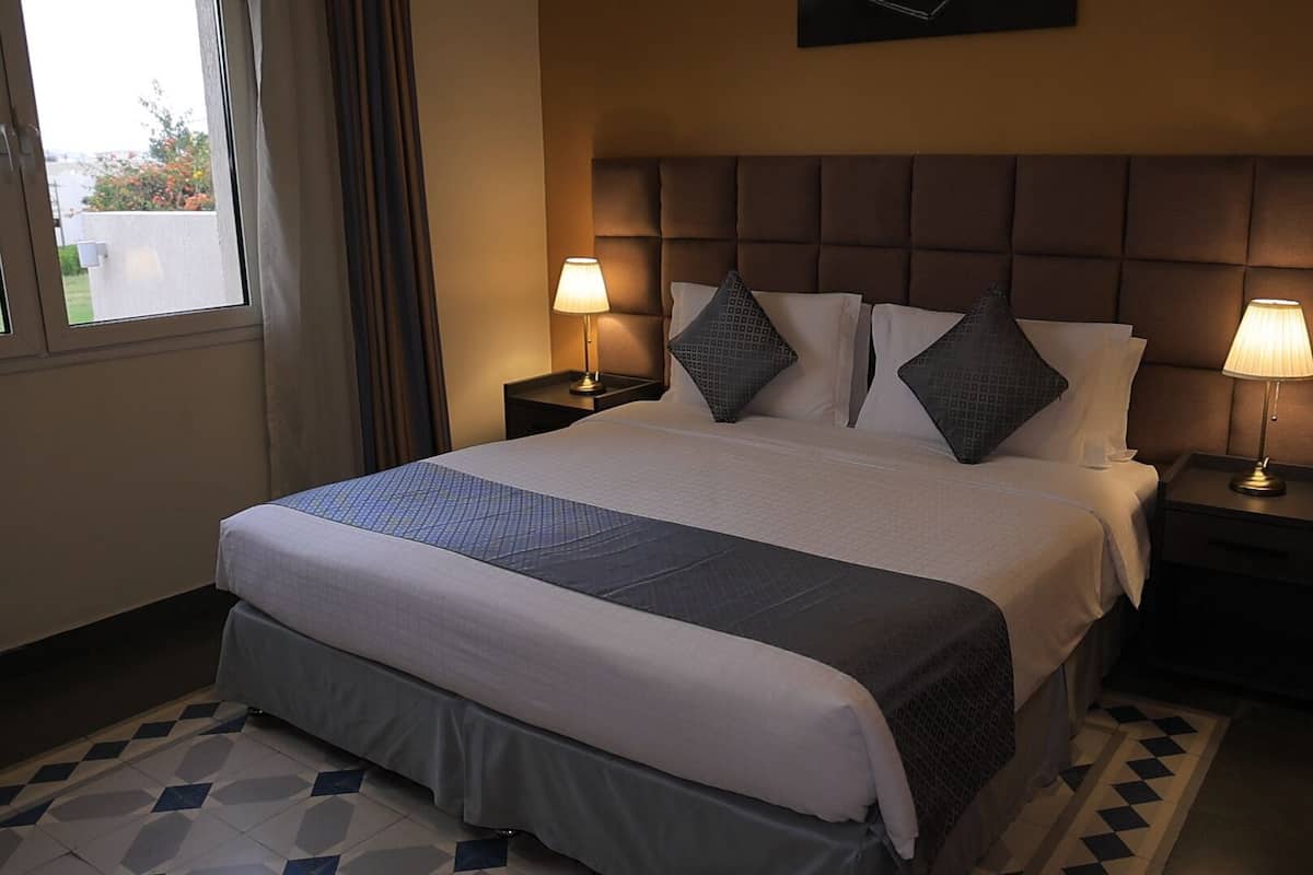 A hotel room with a large bed, two bedside lamps, a headboard, and a window with curtains.