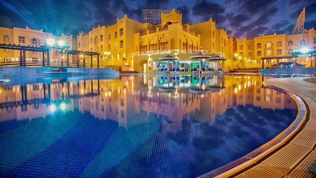 Un hotel con una piscina grande, una zona cubierta con asientos y el reflejo del edificio en el agua.