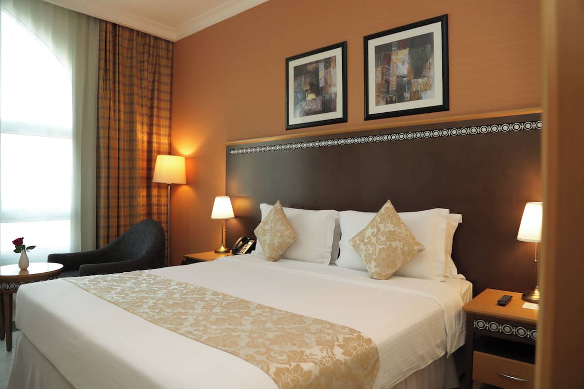 A hotel room with a large bed, two bedside lamps, a chair, a small table with a vase, and two framed pictures on the wall.