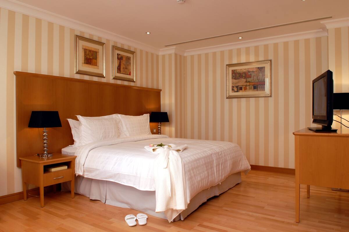A neatly arranged hotel room with a large bed, two bedside tables with lamps, a wooden nightstand, a television on a wooden stand, and striped wallpaper.