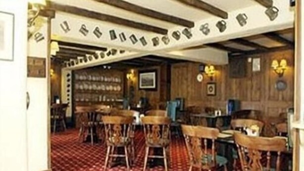 A traditional inn with wooden walls and a patterned carpet. There are several wooden chairs and tables, and a bar area is visible in the background. - The Crown Inn (Swindon)