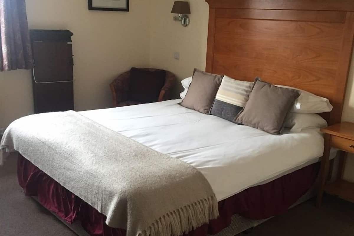 Quarto de hotel com uma cama grande, cabeceira de madeira, mesa de cabeceira, uma cadeira e uma luminária fixada na parede.