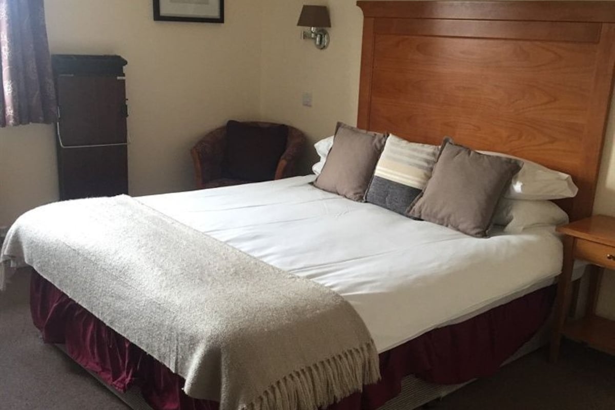 A hotel room with a large bed, a wooden headboard, a nightstand, a chair, and a wall-mounted lamp.