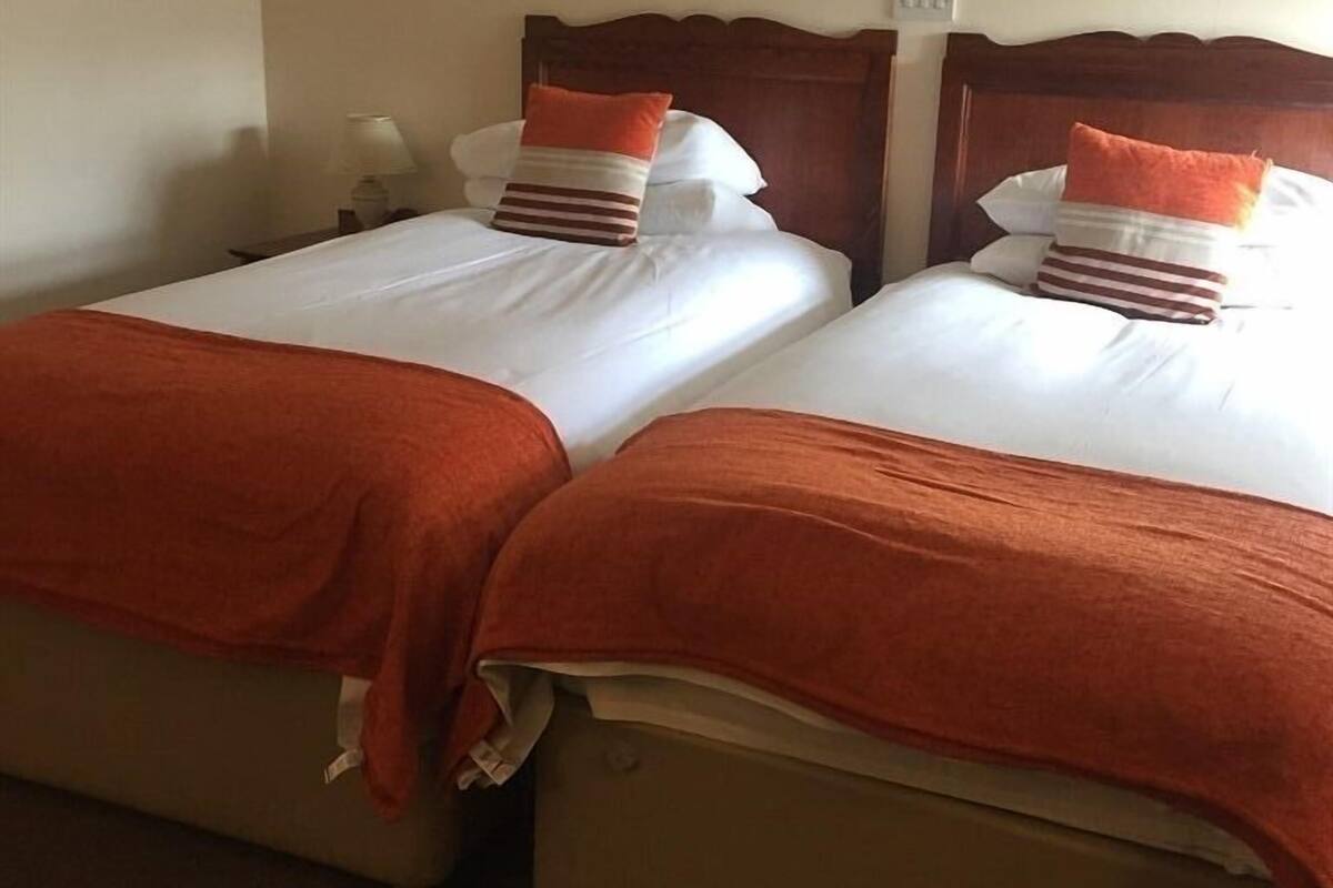 Quarto de hotel com duas camas, cada uma com cabeceira de madeira, roupa de cama branca e almofadas laranja.