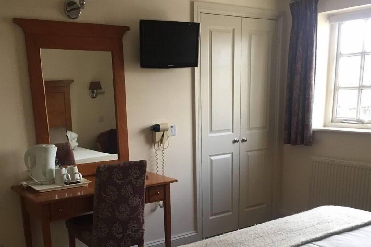 Quarto de hotel com cama, escrivaninha com cadeira, televisão, espelho, porta e janela com cortinas.