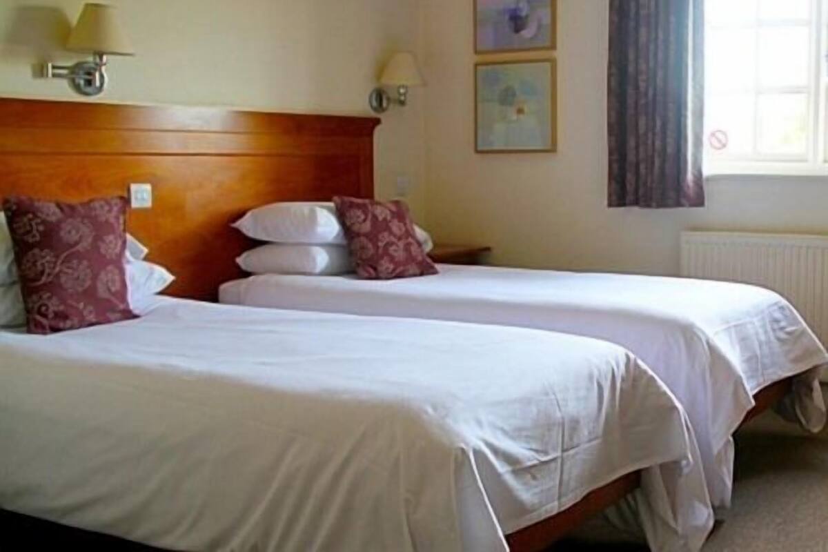 Quarto de hotel com duas camas, cada uma com roupa de cama branca e várias almofadas. Há luminárias na parede e duas fotografias emolduradas.