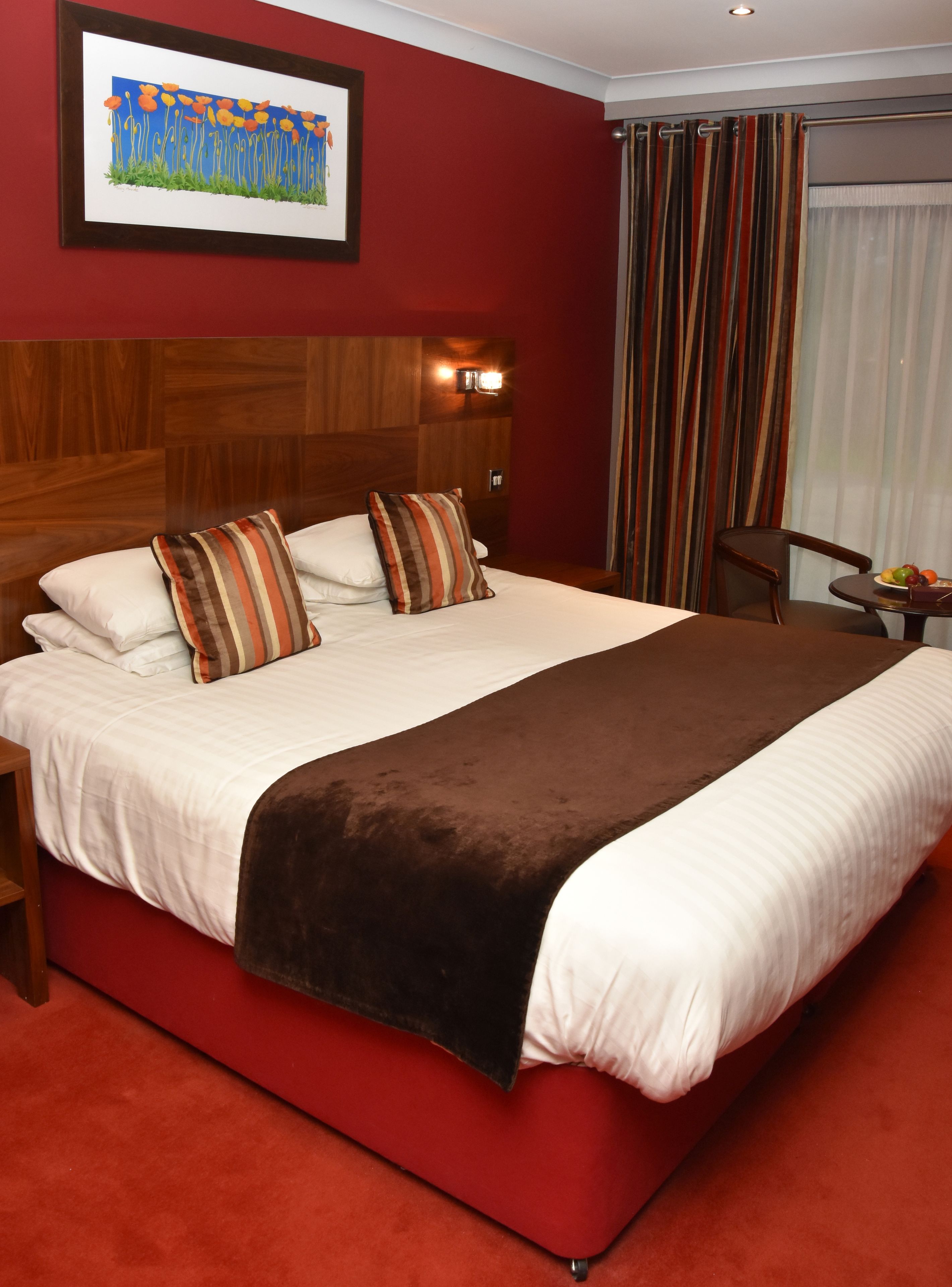 A large bed with multiple pillows, a wooden headboard, a red carpet, a small table with fruit, and a painting on the wall.