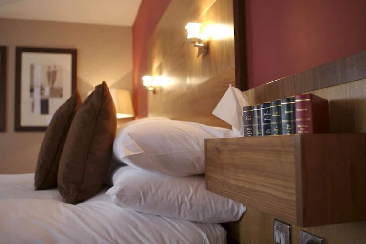 Een hotelkamer met een bed, kussens, een houten hoofdbord en een plank met boeken.