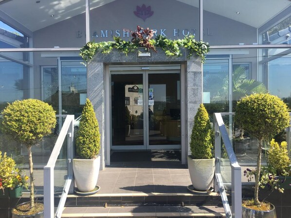 La entrada del Hotel Balmish, con fachada de vidrio moderna, un arco de bienvenida y jardines simétricos.
